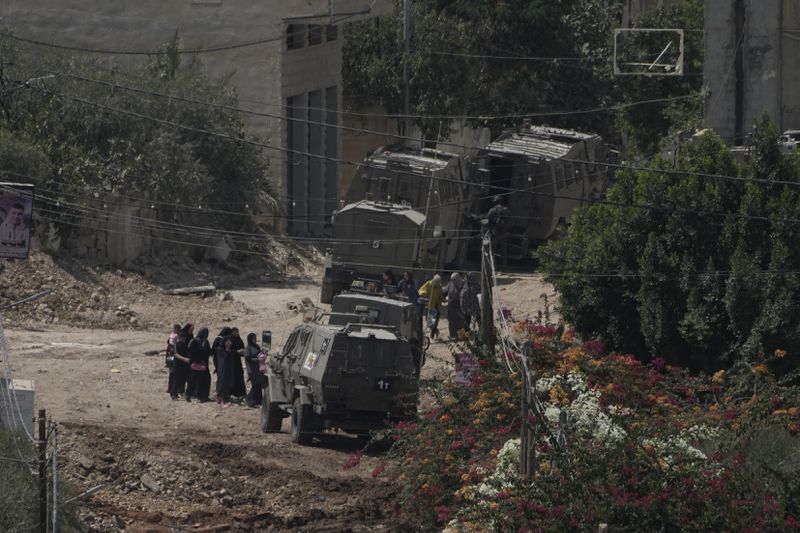 Palestinians stand in line next to Israeli armoured vehicles during a military operation in the West Bank Jenin refugee camp, Saturday, Aug. 31, 2024. (AP Photo/Majdi Mohammed)