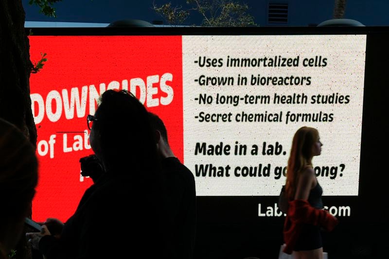 People stand outside the entrance to a pop-up tasting event for "lab-grown" meat produced by Upside Foods, as a protest truck parked out front displays a message advocating against cultivated meat, Thursday, June 27, 2024, in Miami. As Florida's ban on lab-grown meat is set to go into effect next week, one manufacturer hosted a tasting party, serving up cultivated chicken tostadas to dozens of attendees on a rooftop in Miami's Wynwood neighborhood. (AP Photo/Rebecca Blackwell)