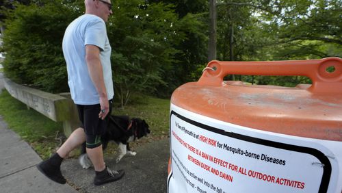 A passer-by walks a dog, Monday, Aug. 26, 2024, while entering a walkway, in Plymouth, Mass., near a sign that advises people of a ban in effect for outdoor activity between dusk and dawn due to the risk of exposure to mosquito-borne diseases. (AP Photo/Steven Senne)