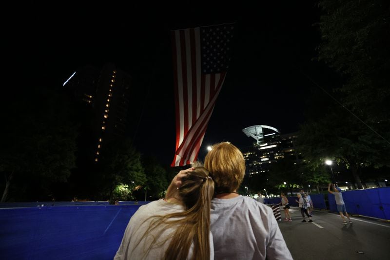 Sally Love Cannaloy and Megan Lienau embrace as they watch the rise of the flag at the starting line of the 55th running of the Atlanta Journal-Constitution Peachtree Road Race in Atlanta on Thursday, July 4, 2024.   (Miguel Martinez / AJC)