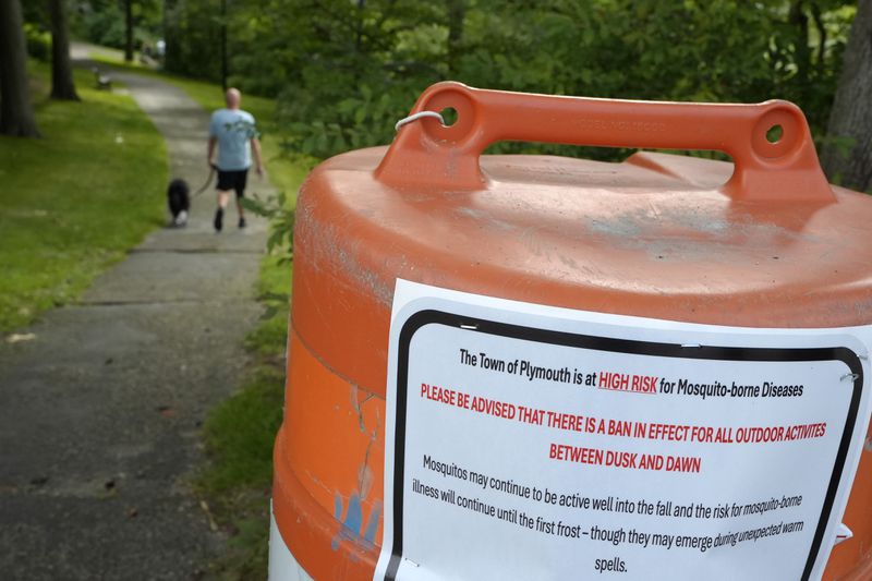 A passer-by walks a dog, Monday, Aug. 26, 2024, along a walkway, in Plymouth, Mass., near a sign that advises people of a ban in effect for outdoor activity between dusk and dawn due to the risk of exposure to mosquito-borne diseases. (AP Photo/Steven Senne)