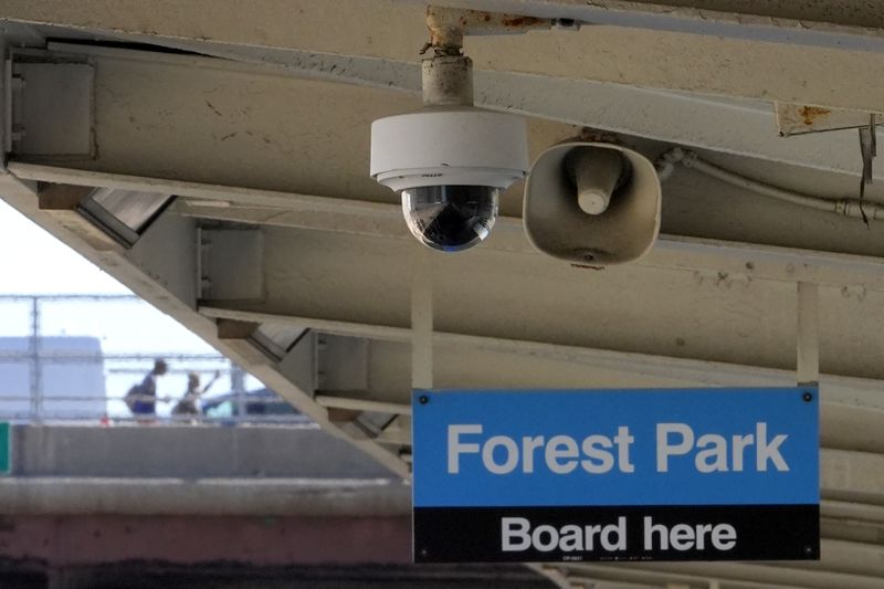 A security camera and speaker hang from the ceiling of the Chicago Transit Authority Harlem Ave. station over the rails that head West to Forest Park, Ill., station as two pedestrians walk toward the station, Tuesday, Sept. 3, 2024, in Forest Park, Ill. (AP Photo/Charles Rex Arbogast)