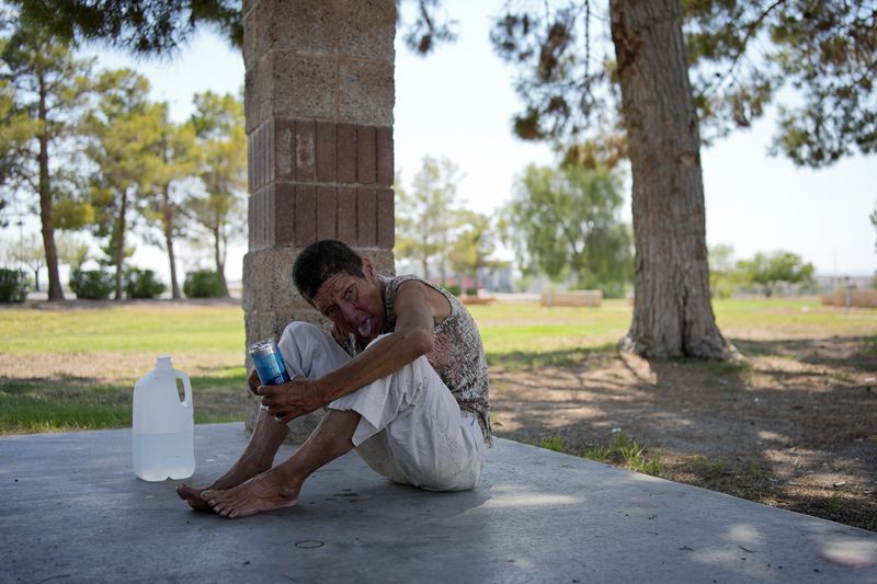 Deb Billet, 66, rests on the ground while drinking cold water given to her by Henderson Public Response officers before they called an ambulance to take her to hospital for heat-related symptoms Wednesday, July 10, 2024, in Henderson, Nev. Billet has been living on the street. About 14 officers from the Office of Public Response in Henderson drove around the city Wednesday, offering water, electrolytes, free bus tickets, and rides to cooling centers during a heat emergency. (AP Photo/John Locher)