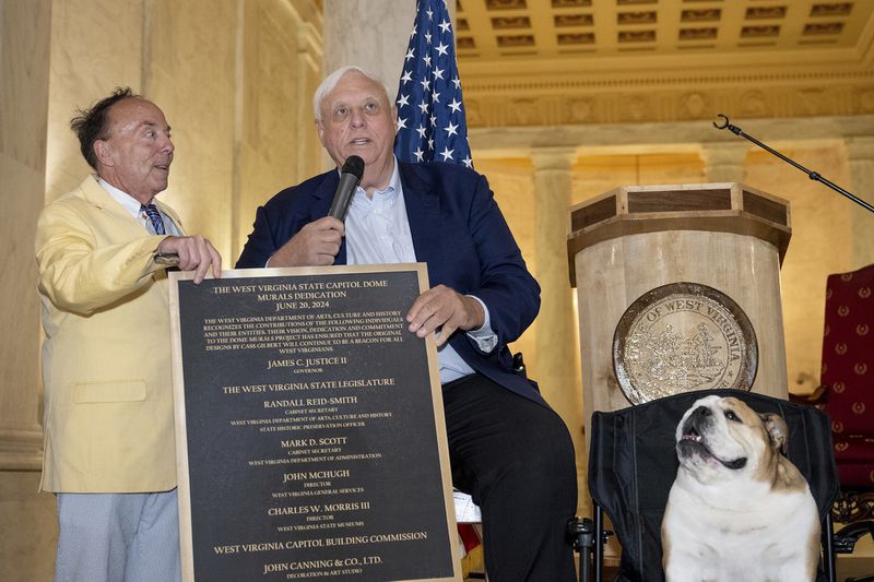 This photo provided by the West Virginia Legislature shows Randall Reid-Smith, West Virginia secretary of the Department of Arts, Culture and History, left, as he hands Gov. Jim Justice, center, a commemorative plaque about four new murals installed at the West Virginia Capitol in Charleston, W.Va., Thursday, June 20, 2024. (Perry Bennett/West Virginia Legislature via AP)