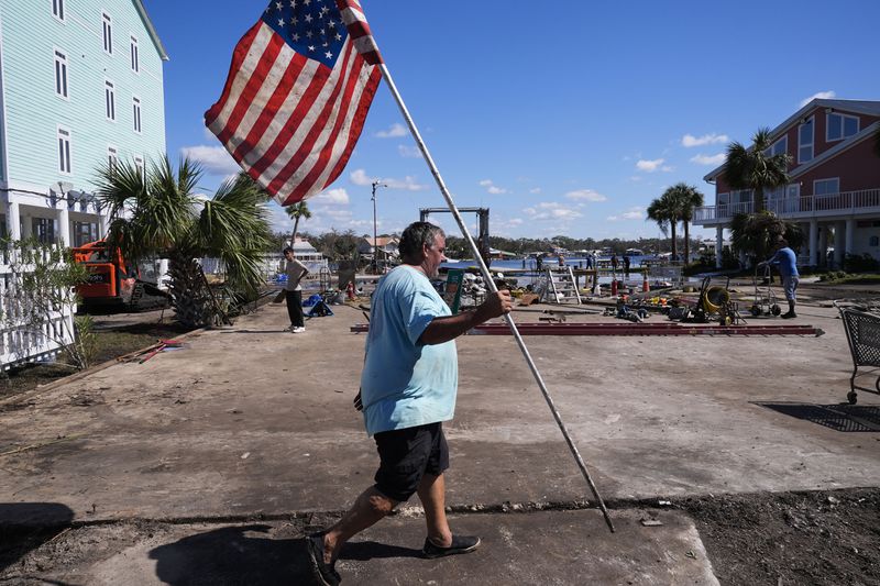 Daniel Dickert walks to plant an American flag on is property were his boat shed was destroyed and his home damaged in the aftermath of Hurricane Helene, in Jena, Fla., Sunday, Sept. 29, 2024. (AP Photo/Gerald Herbert)