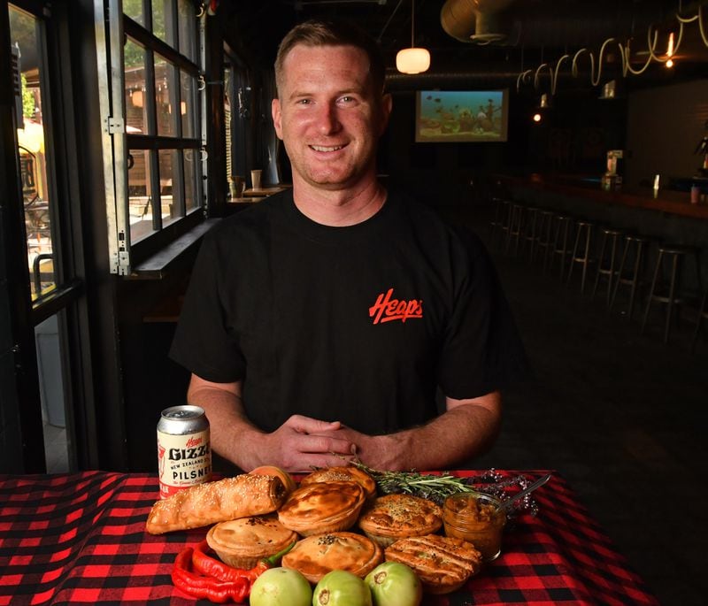 230818 Atlanta, Ga: Heaps Atlanta owner, Jake Harvey, with a few of the products he offers, including (in no particular order): Pork & Sage Salsa Roll, Carnitas Al Pastor Pie, Chana Masala Pie, Lamb & Vegetable Pie, Chicken Pot Pie, Green Tomato Pie, Beef & Cheddar Pie, photographed with the award winning Heaps Gizza - New Zealand Style Pilsner. The AJC visits chef Jake Harvey at his business, Heaps Atlanta, in the Boggs Social building at 1310 White Street SW 30310 in Atlanta, Ga. PHOTO FOR FOOD COVER STORY ON NEW ZEALAND MEAT PIES AAJC090623MEATPIES (CHRIS HUNT FOR THE ATLANTA JOURNAL-CONSTITUTION)