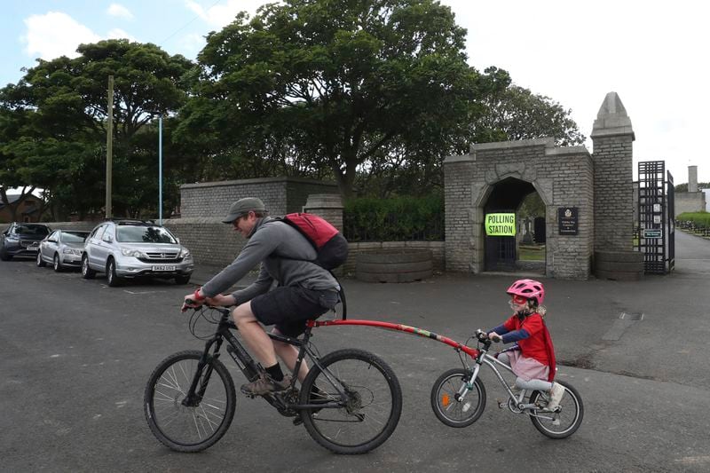 A man cycles with his daughter after casting his vote at Whitley Bay cemetery where there is polling station inside, in Whitley Bay, England, Thursday, July 4, 2024. Britain goes to the polls Thursday after Prime Minister Rishi Sunak called a general election. (AP Photo/Scott Heppell)