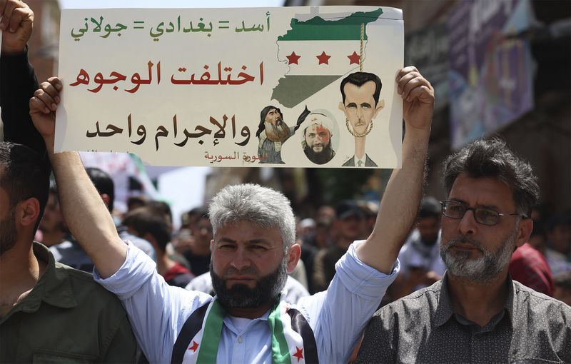 FILE - A Syrian protester holds up a placard during a protest against al-Qaida-linked Hayat Tahrir al-Sham group in Binnish town, in Idlib province, Syria, on May 17, 2024. Syria's devastating civil war, now in its 14th year, remains largely frozen and so are efforts to find a viable political solution to end it, while millions of Syrians have been pulled into poverty, and struggle with accessing food and health care as the economy deteriorates across the country's front lines. Aid organizations are making their annual pitches to donors ahead of a fundraising conference in Brussels for Syria on Monday, May 27. The placard shows drawings of Syrian President Bashar Assad, the late Islamic State group leader Abu Bakr al-Baghdadi and Abu Mohammed al-Golani, leader of the Hayat Tahrir al-Sham, once linked to al-Qaida. The Arabic writing reads: "Assad = Baghdadi = al-Golani. The faces are different but the crime is one." (AP Photo/Ghaith Alsayed, File)