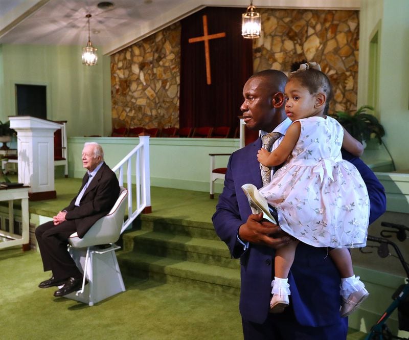 The Rev. Tony Lowden holds his daughter, Tabitha, as he stands in the sanctuary of Maranatha Baptist Church in Plains as he and former President Jimmy Carter, seated, wait to greet visitors following a worship service in June 2019. Lowden, who came to the church earlier that year has formed a tight bond with Carter. “People who try to define him by politics miss the boat completely,” Lowden said. “I’ve had the opportunity to meet with presidents and governors and religious leaders across the nation. But I’ve never met anyone like Jimmy Carter.” Curtis Compton/ccompton@ajc.com