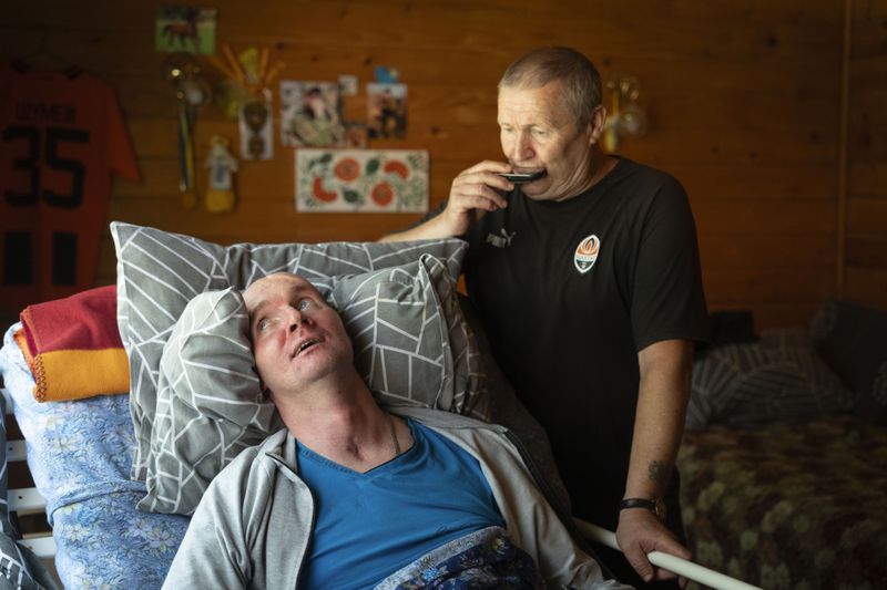 Serhii Shumei, 65, plays harmonica to his son Vitalii, 36, a Ukrainian officer heavily wounded in a battle with Russian forces, at the rehabilitation centre in Modrychi, Western Ukraine, Wednesday, Aug. 28, 2024. (AP Photo/Efrem Lukatsky)