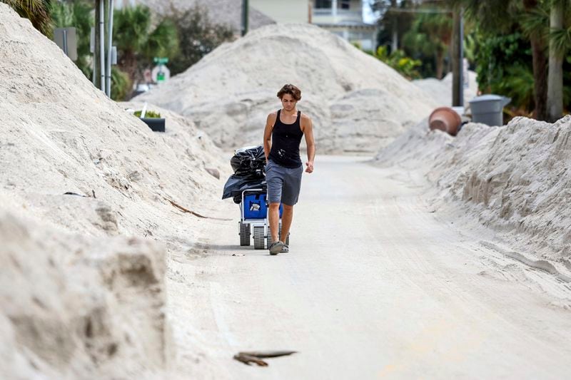 David DeMeza walks out with belongings through sands pushed on to the streets by Hurricane Helene, Wednesday, Oct. 2, 2024, in Treasure Island, Fla. (AP Photo/Mike Carlson)