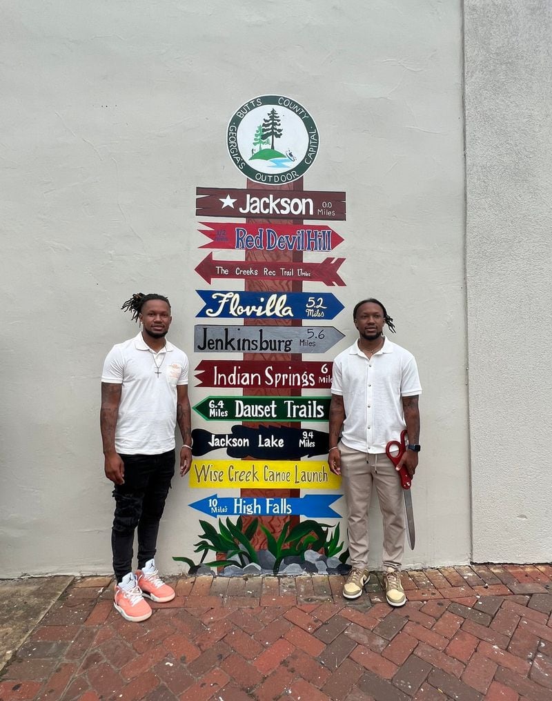 Local artists Chanse Taylor and Chase Taylor of ArtsbyChanse&Chase, LLC stand by the directional sign mural they created just off the Jackson square. (Photo Courtesy of Butts County Chamber of Commerce)