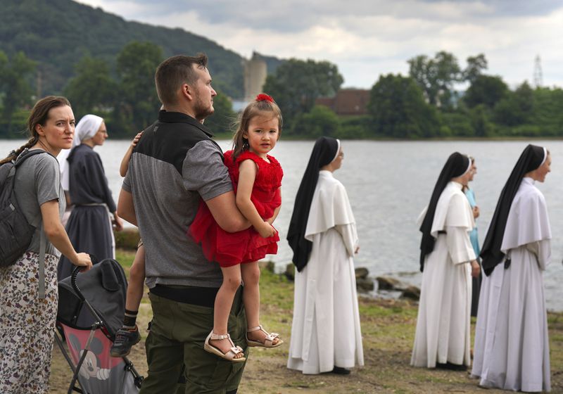 Amanda and Toby Byrnes of Chester, W.Va., stand along the Ohio River with their children and fellow Catholics watching as a boat holding the Eucharist floats toward Wheeling, West Virginia, Sunday June 23, 2024, in Steubenville, Ohio. (AP Photo/Jessie Wardarski)