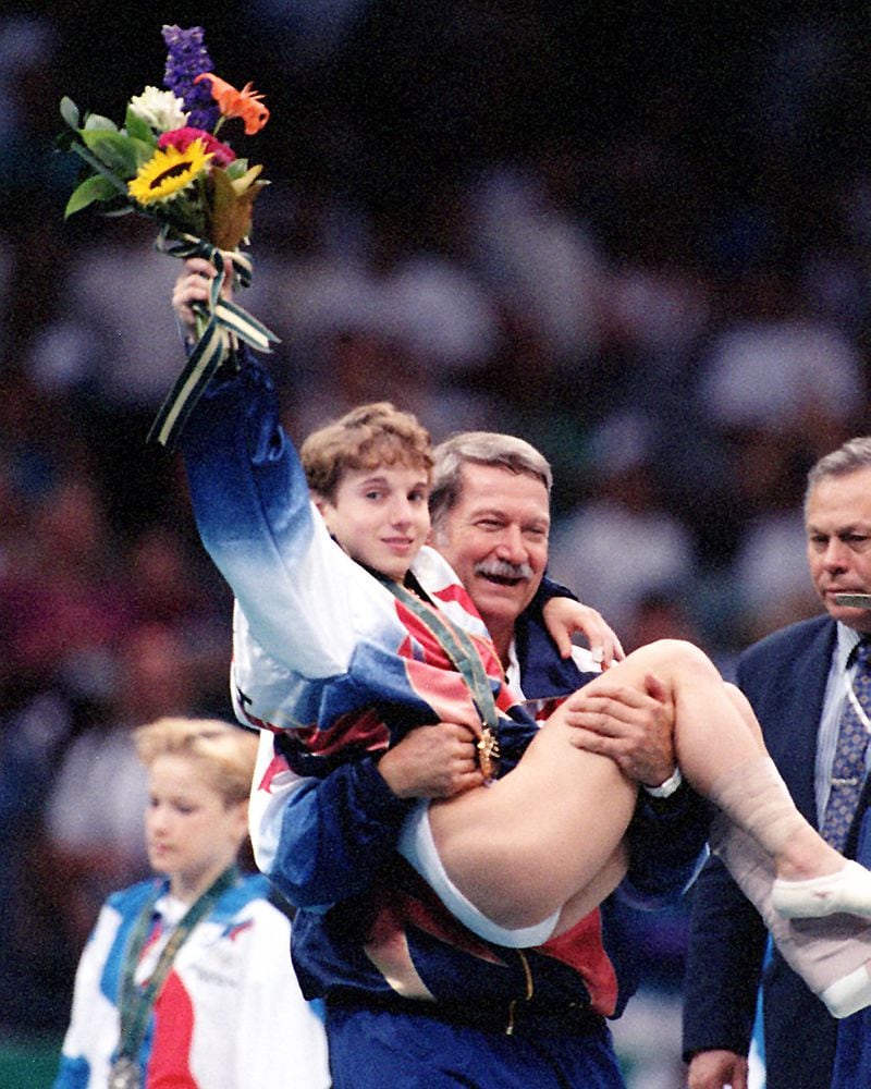 Kerri Strug hoists her flowers to the cheering crowd as she is carried from the podium by team coach, Bela Karolyi at the Georgia Dome on Tuesday, July 23, 1996, during the 1996 Summer Olympic Games in Atlanta, Georgia. Strug was a member U.S. women's gymnastics team that captured the gold medal. She was injured in the final event, her second vault. (Skip Peterson/AJC)