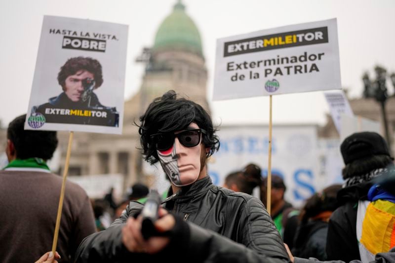 A masked anti-government protester aims a toy gun during a demonstration outside Congress where lawmakers debate a reform bill promoted by President Javier Milei in Buenos Aires, Argentina, Wednesday, June 12, 2024. The sign at right reads in Spanish "Exterminator of the homeland." (AP Photo/Natacha Pisarenko)