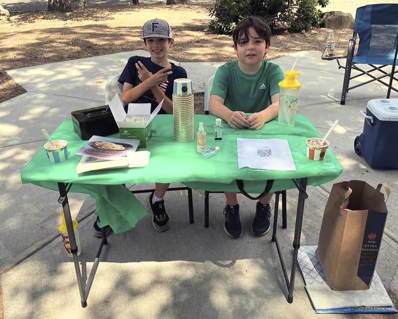 In this Saturday, Aug. 3, 2024 photo provided by Nancy Doherty, Ben Doherty, of Braintree, Mass., left, and his cousin Danny Doherty, 12, of Norwood, Mass., right, sit at a homemade ice cream stand near Danny's home in Norwood. (Nancy Doherty via AP)