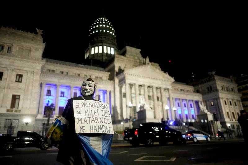 A demonstrator holds a banner that reads in Spanish "The 2025 budget, starving us to death" as Argentina's President Javier Milei presents next year's budget in Congress in Buenos Aires, Argentina, Sunday, Sept. 15, 2024. (AP Photo/Natacha Pisarenko)