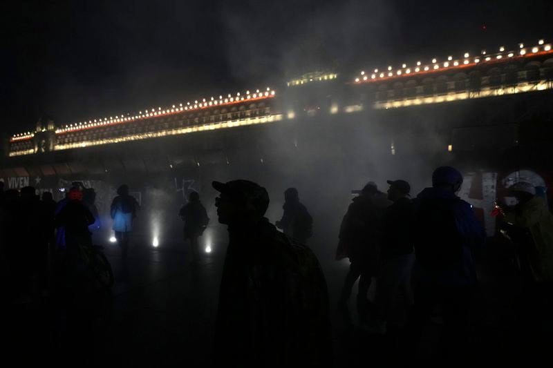 Youths are silhouetted in clouds of smoke caused by fire balls thrown at the barriers protecting the facade of the National Palace during a demonstration marking the 10-year anniversary of the disappearance of 43 students from an Ayotzinapa rural teacher's college, in Mexico City, Thursday, Sept. 26, 2024. (AP Photo/Fernando Llano)