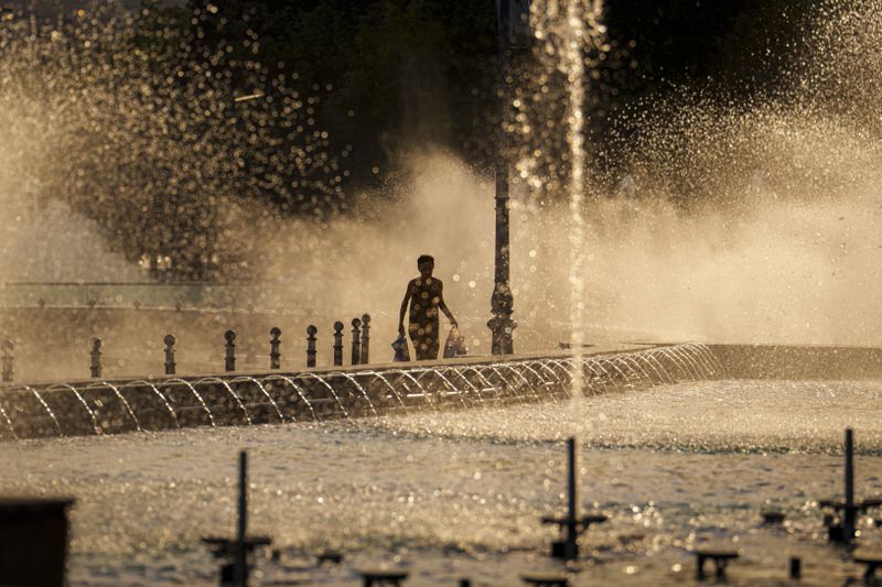 A woman carries plastic bags walking by a public fountain before sunset in Bucharest, Romania, Thursday, June 20, 2024 as temperatures exceeded 38 degrees Celsius (100.4 Fahrenheit). The national weather forecaster issued a orange warning for western and southern Romania where temperatures are expected to reach 38 degrees Celsius (100.4 Fahrenheit) in the coming days. (AP Photo/Vadim Ghirda)