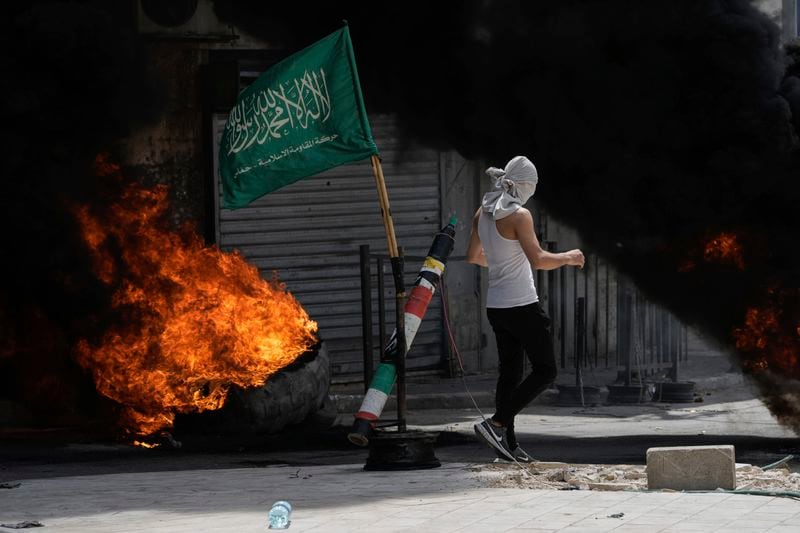 A masked Palestinian demonstrator burns tires next to a mock rocket and the Hamas militant group flag during clashes with Israeli forces in the West Bank city of Jenin, Wednesday, May 22, 2024. (AP Photo/Majdi Mohammed)