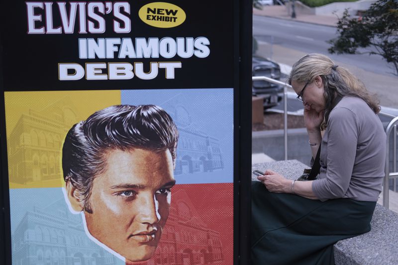 Sandra Morris sits outside the Ryman plaza next to a poster of commemorating a performance by Elvis Presley at the concert venue in Nashville, Tenn., on July 30, 2024. (AP Photo/Luis Andres Henao)