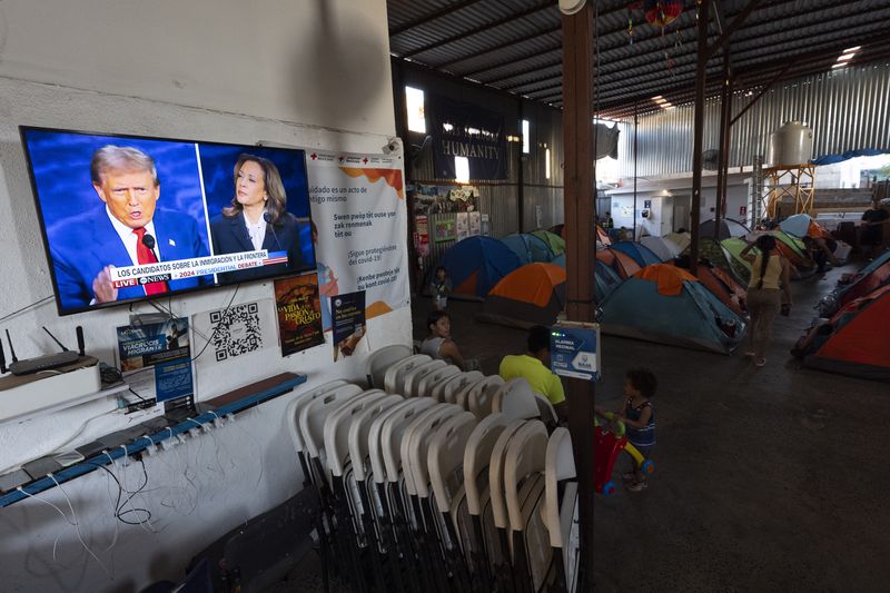 An asylum-seeking migrant from Michoacan, Mexico, walks back to the tent she shares with her family as the presidential debate between Republican presidential nominee former President Donald Trump and Democratic presidential nominee Vice President Kamala Harris plays on a television Tuesday, Sept. 10, 2024, at a shelter for migrants in Tijuana, Mexico. (AP Photo/Gregory Bull)