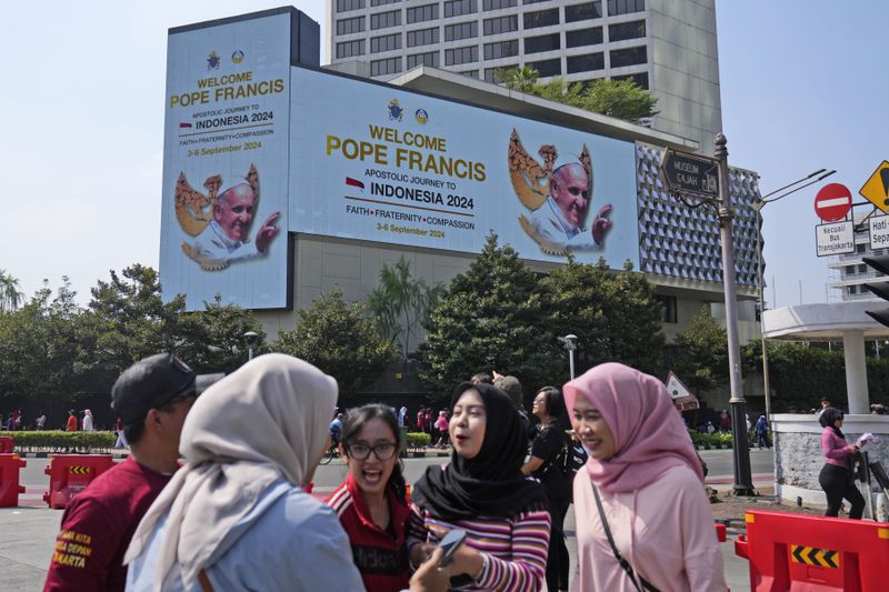 Muslim women chat as a large digital advertisement board with a welcoming message for Pope Francis is displayed on the facade of a building nearby in Jakarta, Sunday, Sept. 1, 2024, ahead of his visit to Indonesia from Sept. 3-6. (AP Photo/Dita Alangkara)