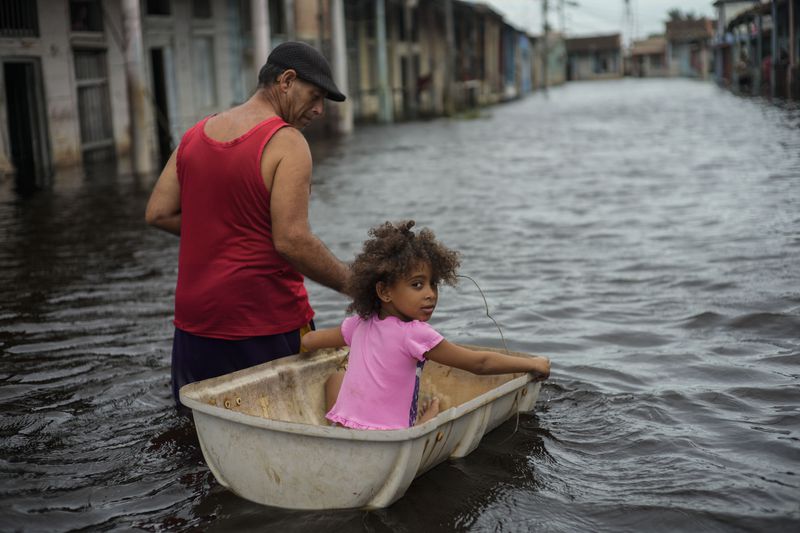 Jesus Hernandez guides his granddaughter Angelina via a container through a street flooded in the passing of Hurricane Helene, in Batabano, Mayabeque province, Cuba, Thursday, Sept. 26, 2024. (AP Photo/Ramon Espinosa)