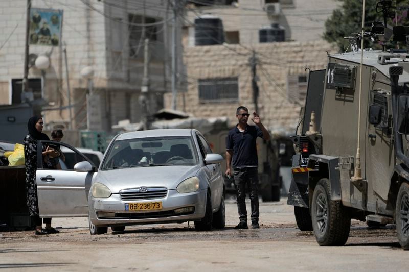 A man waves to members of the Israeli forces inside an armoured vehicle, as he stands next to his car with a woman holding a baby during a military operation in the West Bank city of Jenin, Wednesday, Aug. 28, 2024. (AP Photo/Majdi Mohammed)