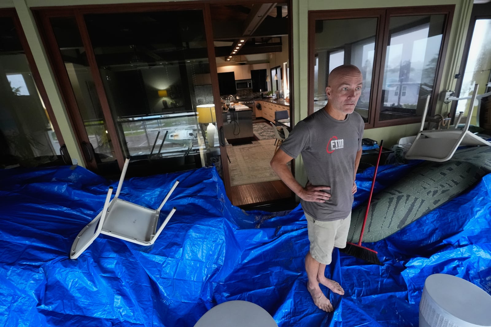 Christian Burke stands on the third floor of his home, where he, his mother, and his aunt plan to ride out Hurricane Milton with views through hurricane proof glass over Tampa Bay, in Gulfport, Fla., Wednesday, Oct. 9, 2024. Burke, who said his engineer father built the concrete home to withstand a Category 5 hurricane, expects his home to turn into an island, with up to 8 feet of water filling the raised first floor. Behind Burke, a window reflects a boat deposited by Hurricane Helene in the bay front park across the street. (AP Photo/Rebecca Blackwell)