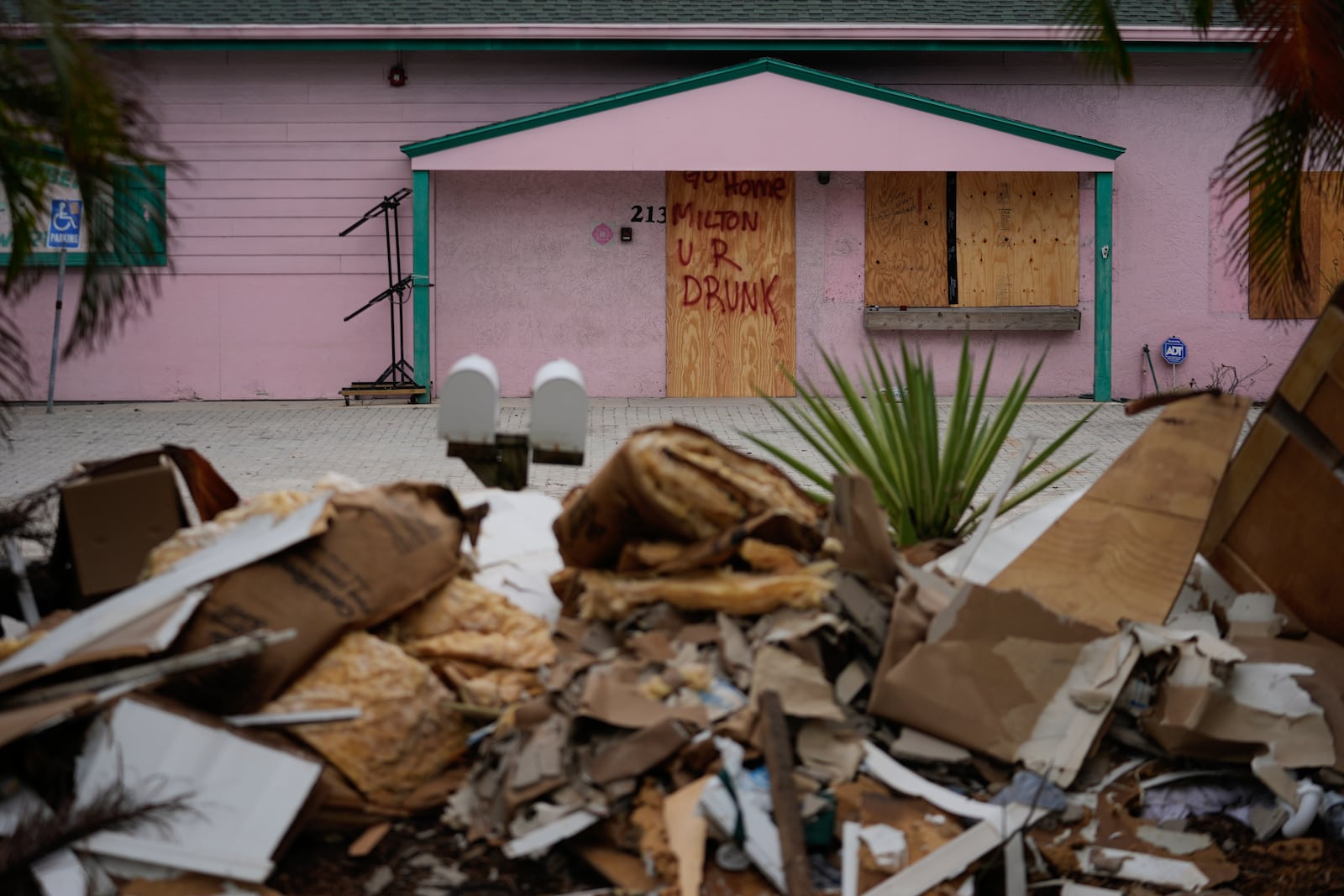 A boarded up business, marked with graffiti reading "Go home Milton, U R drunk," is seen past debris from Hurricane Helene flooding piled up outside a home, ahead of the arrival of Hurricane Milton, in Holmes Beach on Anna Maria Island, Fla., Tuesday, Oct. 8, 2024. (AP Photo/Rebecca Blackwell)