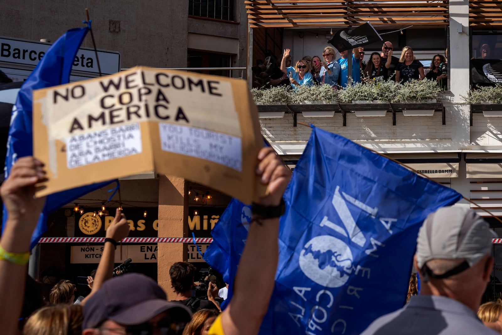 Supporters of America's Cup wave from a restaurant as demonstrators march shouting slogans against the holding of the America's Cup sailing competition, during a protest demanding the right to housing, in Barcelona, Spain, Sunday, Oct. 13, 2024. (AP Photo/Emilio Morenatti)