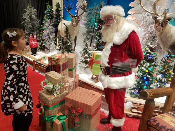 Visits to Santa at metro Atlanta malls and stores during the pandemic are often still happening, but with a slate of new precautions in place. Props kept nine-year-old Abby Rollins separated from Santa, who wore a face shield, on a recent visit at the Bass Pro Shops store in Duluth. MATT KEMPNER / AJC