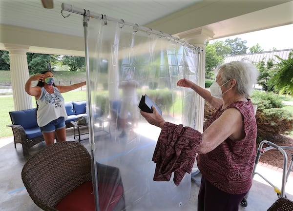 Elsie Grant, 86, sets her walker aside reaching for an air hug from her daughter Wanda Schroeder, socially distanced behind a protective plastic curtain when families could visit loved ones in the outdoor pavilion at Westbury Medical Care & Rehab earlier this month. CURTIS COMPTON / CCOMPTON@AJC.COM