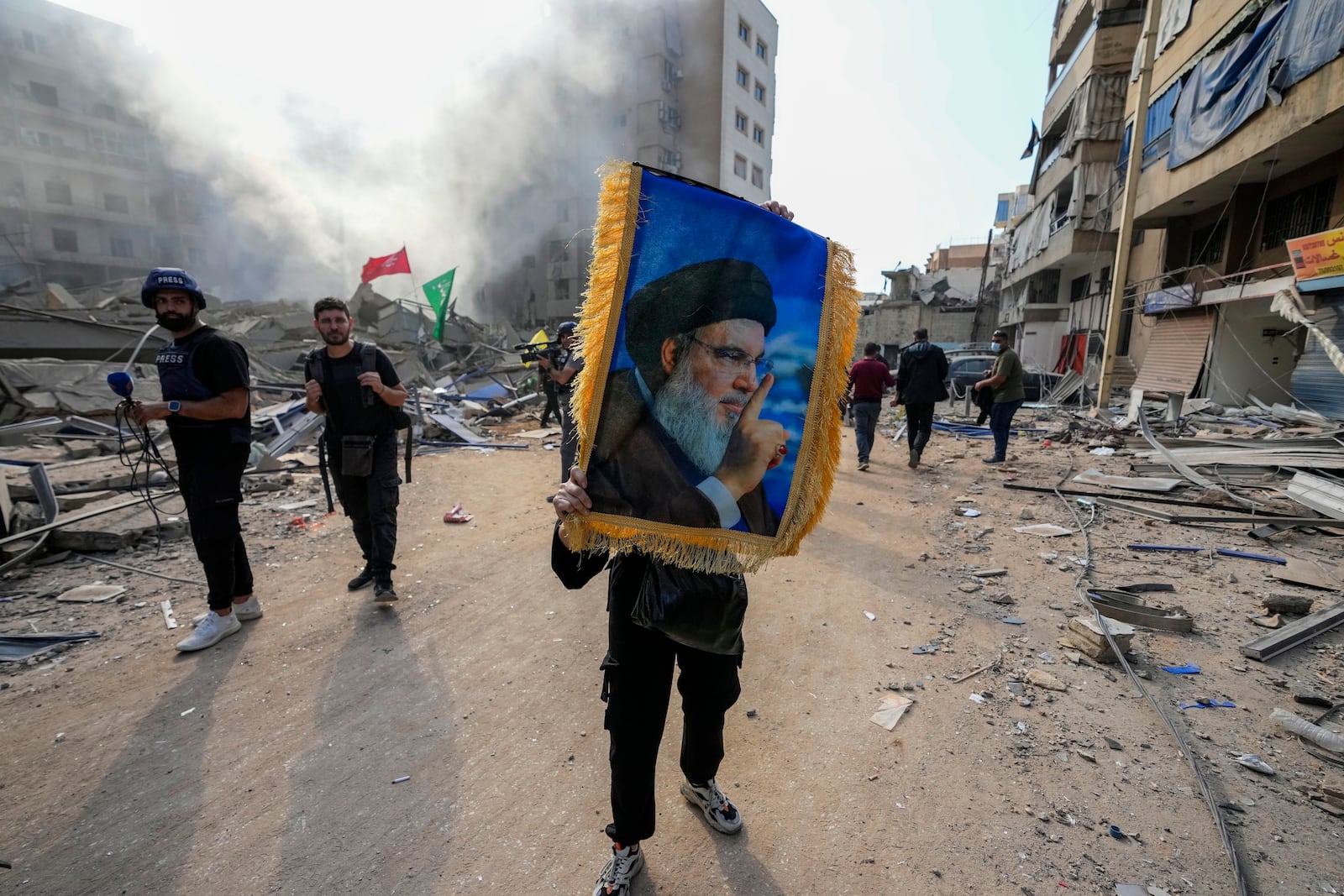 A woman holds a picture of slain Hezbollah leader Hassan Nasrallah as smoke rises from the site of an Israeli airstrike in Dahiyeh, Beirut, Lebanon, Friday, Nov. 1, 2024. (AP Photo/Hassan Ammar)