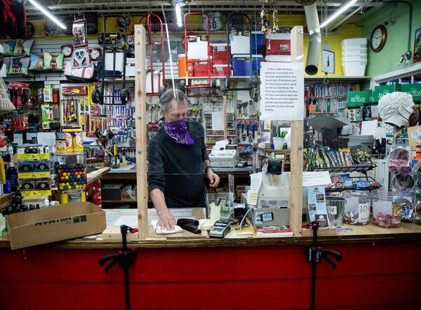 Intown Ace Hardware owner Doug Eifrid sanitizes the counters between customers while standing behind a temporary plexiglass shield in his store Sunday, April 19, 2020. STEVE SCHAEFER / SPECIAL TO THE AJC