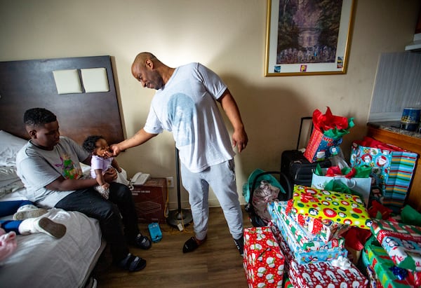 Money White, 13, from left, holds his 5-month-old sister, Justice, while his father, Melvin White, loves on the baby in their extended stay hotel room where Raneice and Melvin live with 5 of their children Dec 21, 2022.  The family has lived in extended stay hotels for years and, despite Melvin working full time, they cannot find long-term, safe housing they can afford to live in. Legal Aid and law firm Lueder, Larkin and Hunter gave the family all their Christmas gifts. Jenni Girtman for the AJC 2022