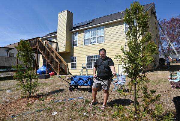 Alex Betancourt is watering a newly planted tree as solar panels on the roof are shown in the background at his home on Friday, March 24, 2023, in Suwanee. Alex Betancourt put solar panels on his roof last year. He received letters from his HOA claiming the panels were an architectural addition that violated the community's rules. Now, they're threatening to remove the panels from his roof. (Hyosub Shin / Hyosub.Shin@ajc.com)