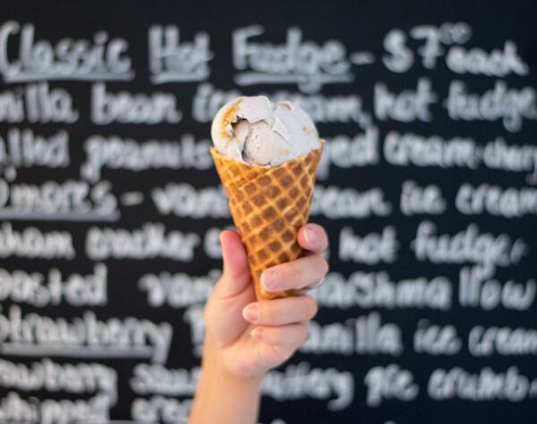 A waffle cone filled with Lavender Honeycomb ice cream is held up in front of the Queen of Cream menu board. CONTRIBUTED BY HENRI HOLLIS