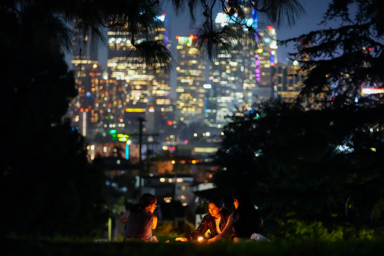 FILE - The downtown Los Angeles skyline gives a backdrop to artists painting under candlelight at Everett Triangle Park, Oct. 22, 2024, in Los Angeles. (AP Photo/Julio Cortez, File)