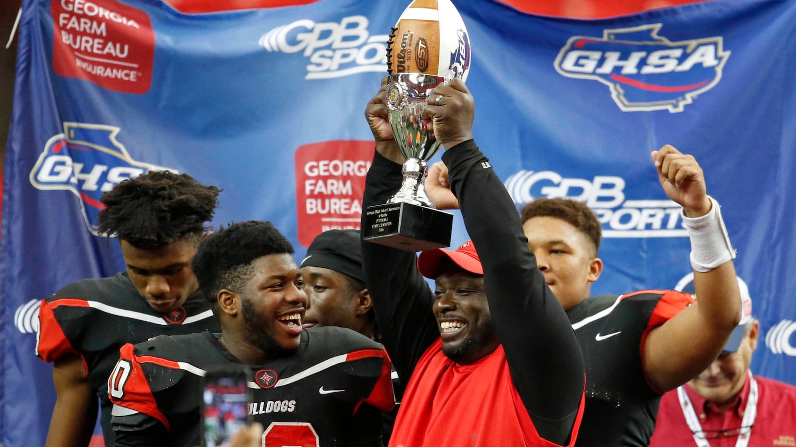 Macon County head coach Dexter Copeland (center) hoists the Class A-Public state championship trophy Saturday, Dec. 10, 2016, at the Georgia Dome in Atlanta. (Jason Getz/Special to AJC)