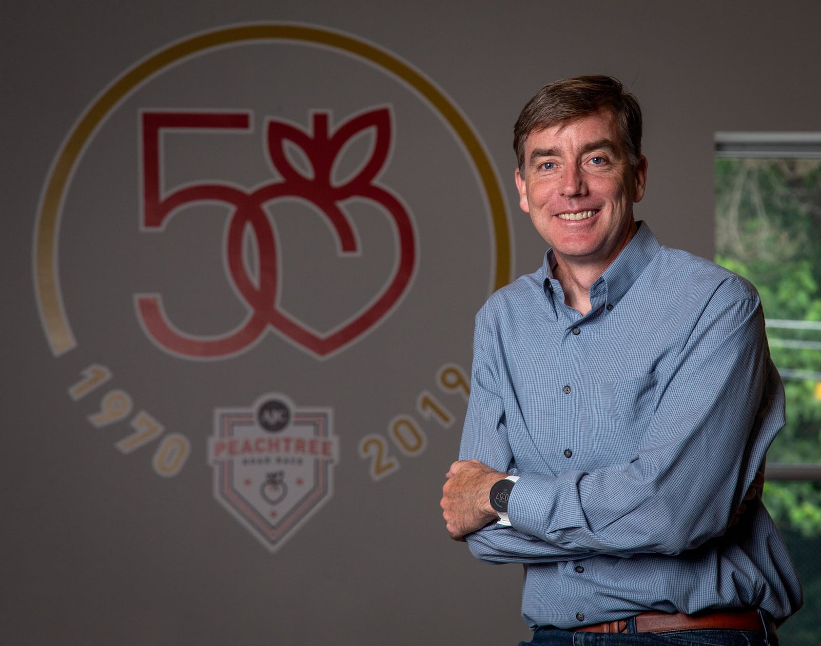 Atlanta track club’s Executive Director Rich Kenah in front of the logo for the 50th Running of the AJC Peachtree Road Race in the track club’s office June 4, 2019. STEVE SCHAEFER / SPECIAL TO THE AJC