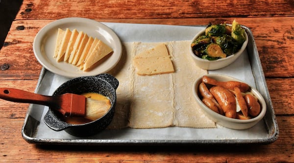 Oven components for the savory puff pastry galette include (clockwise from top left) Thomasville Tomme cheese, Brussels sprouts, fingerling potatoes and egg wash to brush on the puff pastry, all sitting on top of puff pastry dough. (Styling by chef Shain Wancio / Chris Hunt for the AJC)