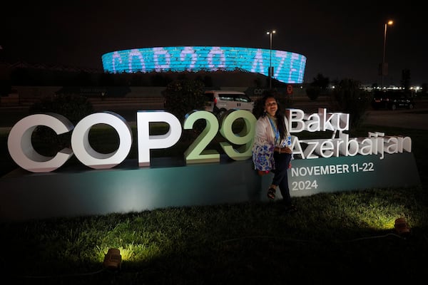 A woman poses for a photo with the Baku Olympic Stadium in the background as it is illuminated at the COP29 U.N. Climate Summit, Monday, Nov. 11, 2024, in Baku, Azerbaijan. (AP Photo/Peter Dejong)