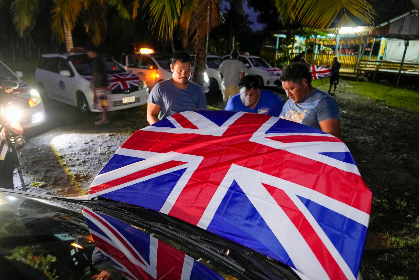 Men attach Union Jacks to the hoods of their taxis as they wait for the arrival of Britain's King Charles III and Queen Camilla in the village of Siumu, Samoa, on Wednesday, Oct. 23, 2024. (AP Photo/Rick Rycroft)