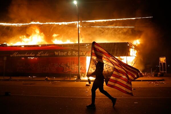 A protester carries a U.S. flag upside, a sign of distress, next to a burning building Thursday, May 28, 2020, in Minneapolis. Protests over the death of George Floyd, a black man who died in police custody Monday, broke out in Minneapolis for a third straight night. (AP Photo/Julio Cortez)