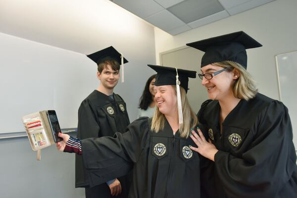 Alex Goodman and Faith Roman (right) pose for a selfie with Kurt Vogel in the background as they try on their caps and gowns ahead of their graduation from the EXCEL program for students with intellectual disabilities at Georgia Tech’s Scheller College of Business. HYOSUB SHIN / HSHIN@AJC.COM