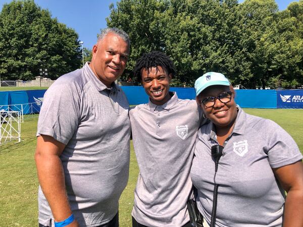 Scott Patton (left) and Linda Hart-Patton (right) and their son Miles Patton (center) pose for a photo in Piedmont Park on July 4, 2021 at the AJC Peachtree Road Race. The three are longtime volunteers for the race, specifically with the pre-race setup of the T-shirt distribution area and the race-day distribution of the shirts. (AJC photo by Ken Sugiura)