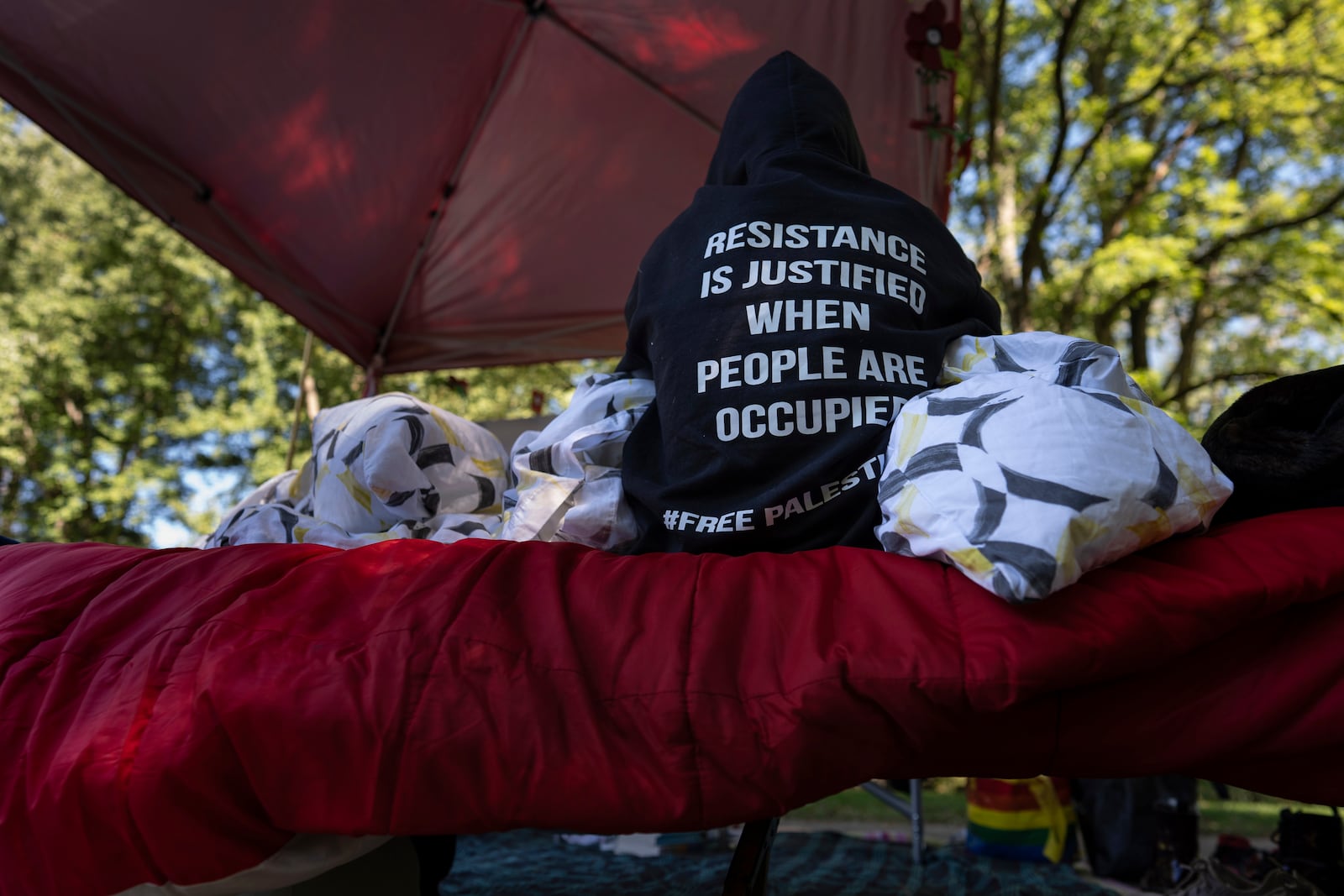 A protester sits on a cot on the side of the road near the driveway of Ohio Democratic Rep. Greg Landsman's Cincinnati residence wearing a sweatshirt that reads "Resistance is justified when people are occupied #Free Palestine," Monday, Oct. 7, 2024. (AP Photo/Carolyn Kaster)