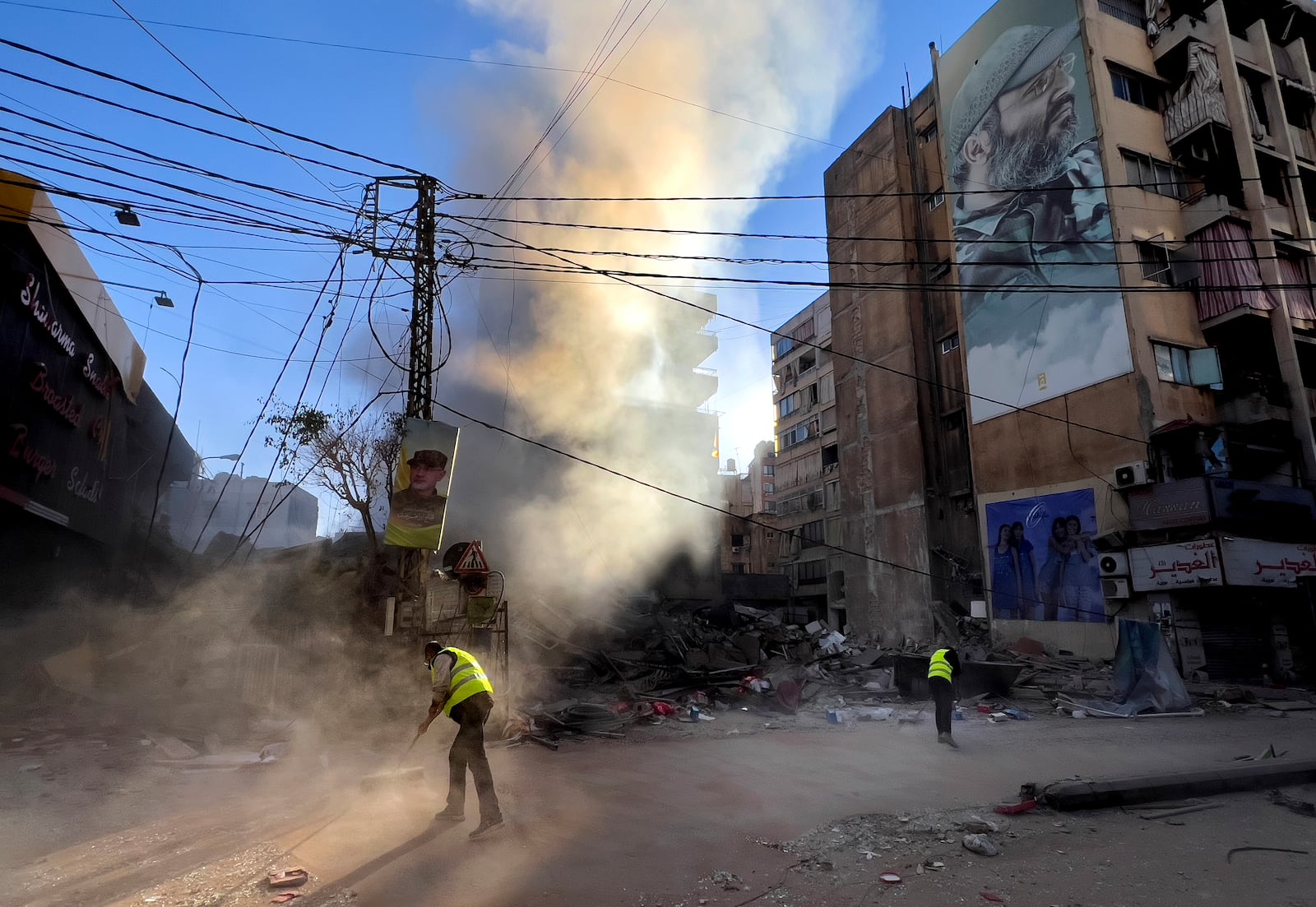 Workers clean a street under a giant portrait of the late Hezbollah military commander Imad Mughniyeh, as smoke rises from a destroyed building that was hit by an Israeli airstrike in Dahiyeh, in the southern suburb of Beirut, Lebanon, Sunday, Oct. 20, 2024. (AP Photo/Hussein Malla)